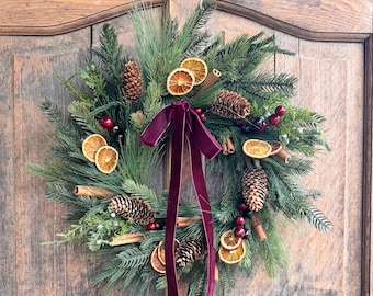 May include: A circular wreath made of green pine branches, decorated with brown pinecones, dried orange slices, cinnamon sticks, and small red berries. A long, deep burgundy velvet ribbon is tied in a bow at the top, hanging down the center.
