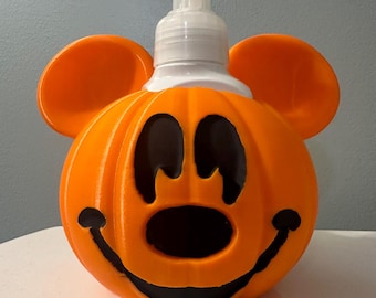May include: Orange pumpkin-shaped soap dispenser with Mickey Mouse ears and a black face. The dispenser has a white foam pump.
