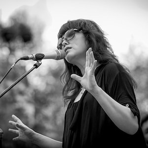 Peut inclure: Une femme aux longs cheveux noirs chante dans un microphone tout en portant des lunettes de soleil et un haut noir.