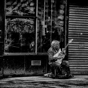 Può includere: Una fotografia in bianco e nero di una persona seduta su un marciapiede che suona la chitarra. La persona indossa una giacca e jeans. La chitarra è di colore chiaro e ha un battipenna nero. La persona è seduta davanti a una vetrina chiusa con un portone scorrevole in metallo.
