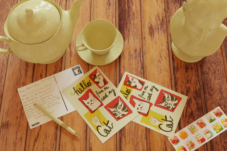 May include: Two postcards with a yellow background and red and black cat illustrations. The postcards say "hello from me and my cat". A handwritten postcard with a message to Mom and Dad. A teapot, teacup, and saucer in a light yellow color. A small stack of vintage stamps with floral designs.