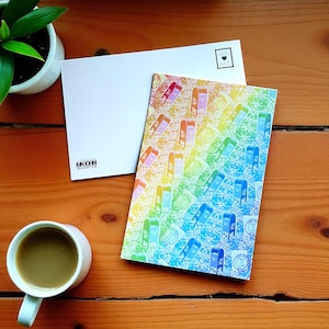 May include: A colorful postcard with a rainbow pattern of stylized arches and stamps. A white envelope is next to the postcard. A cup of coffee and a small potted plant are also on the wooden surface.