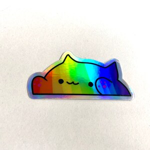 May include: A rainbow holographic sticker of a cat with a paw print on its back. The cat is laying down with its head resting on its paw.