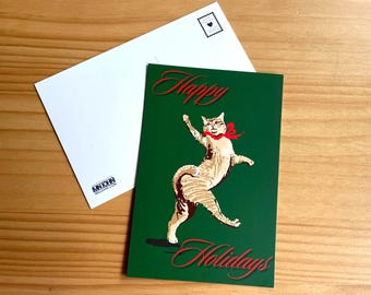 May include: A green holiday greeting card with a dancing cat wearing a red bow, the words "Happy Holidays" in red script. A white envelope is next to the card, with a postage stamp design.