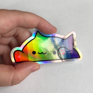 May include: A holographic sticker of a rainbow cat with a cute expression. The cat is lying down with its paws visible. The sticker is made of a shiny, iridescent material.
