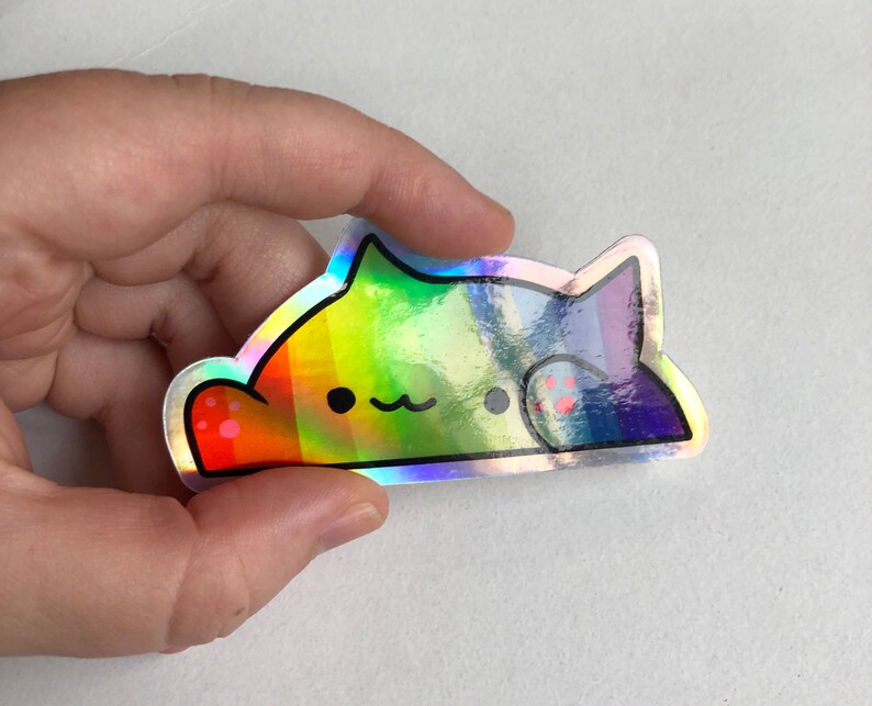 May include: A holographic sticker of a rainbow cat with a cute expression. The cat is lying down with its paws visible. The sticker is made of a shiny, iridescent material.