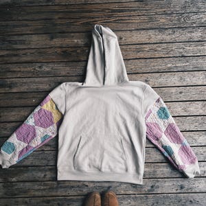 May include: A beige hoodie with a kangaroo pocket and a hood. The sleeves feature a patchwork design with geometric patterns in shades of purple, yellow, and teal. The hoodie is laid out on a wooden surface.