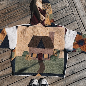 May include: Quilted jacket with a house design. The jacket features a beige background with a brown house, green grass, and a winding path. The hood and sleeves have patchwork patterns in various colors. White sneakers are visible at the bottom.