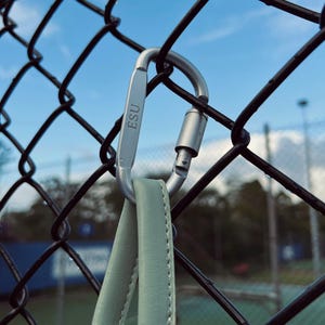 May include: A silver carabiner with the letters "ESU" engraved on it is attached to a black chain-link fence. A light green strap is attached to the carabiner. The background shows a tennis court and a cloudy blue sky.
