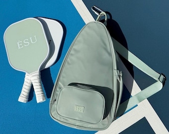 May include: A sage green  paddle bag with a zippered pocket and an adjustable strap. The bag has the word "ESU" embroidered on the front. Two sage green paddles with white handles and the word "ESU" in white lettering are in the background.