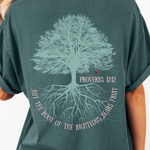 May include: Teal-colored t-shirt featuring a light blue tree graphic on the back. The design includes the text "PROVERBS 12:12" and "BUT THE ROOT OF THE RIGHTEOUS BEARS FRUIT" in a circular arrangement around the tree.