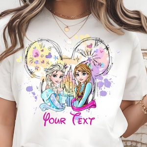 May include: White t-shirt featuring a colorful graphic of two cartoon princesses within a Mickey Mouse ear outline. The design includes the text "Your Text" in pink, with splashes of purple, yellow, and blue.