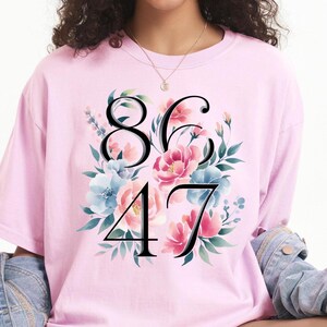 May include: A light pink t-shirt featuring the numbers 86 and 47 in black, surrounded by a floral design with pink and blue flowers and green leaves. The shirt is made of a soft material.