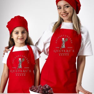 May include: Two people wearing red chef hats and red aprons with the text "Anyone can cook Paris France Gusteau's" and a cartoon rat holding a wooden spoon. The aprons have a pocket on the front. A bowl of red grapes is on a white table.