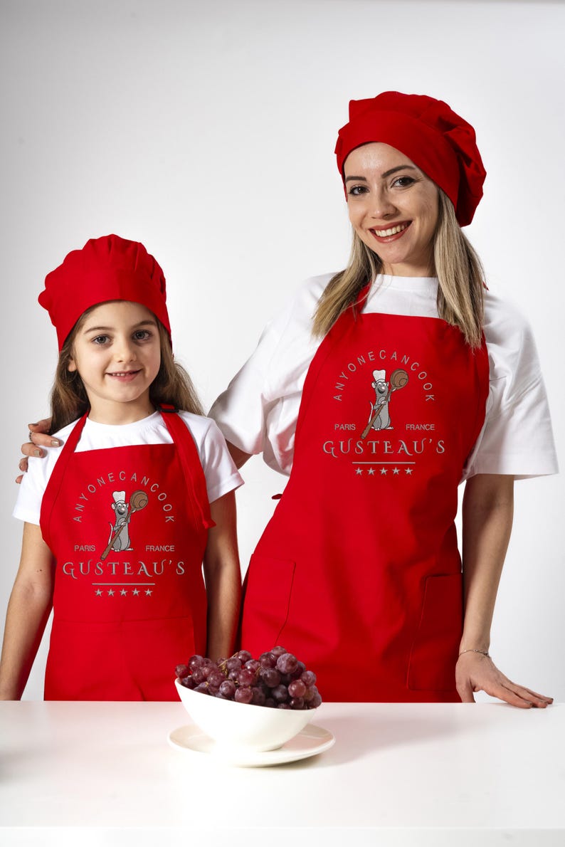 May include: Two people wearing red chef hats and red aprons with the text "Anyone can cook Paris France Gusteau's" and a cartoon rat holding a wooden spoon. The aprons have a pocket on the front. A bowl of red grapes is on a white table.
