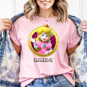 Peut inclure: T-shirt rose avec un dessin de la Princesse Peach dans un cadre dor&eacute; et le nom "EMILY" en dessous. La personne porte une veste en jean avec une doublure &agrave; motifs floraux.