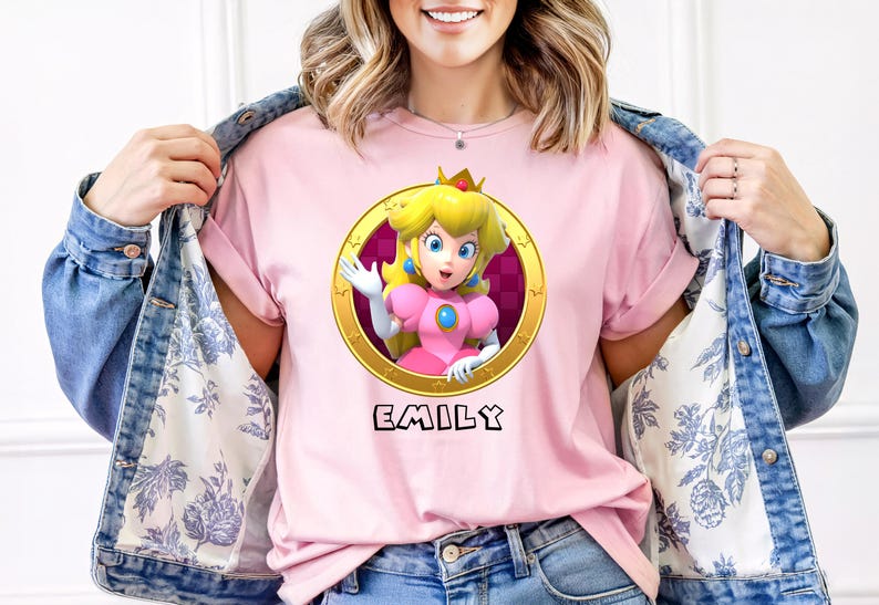 Peut inclure: T-shirt rose avec un dessin de la Princesse Peach dans un cadre dor&eacute; et le nom "EMILY" en dessous. La personne porte une veste en jean avec une doublure &agrave; motifs floraux.