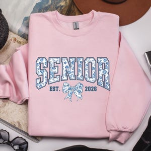 May include: A light pink sweatshirt with the word "SENIOR" in a blue floral design, with "EST. 2026" and a bow graphic. The sweatshirt is a crew neck style.