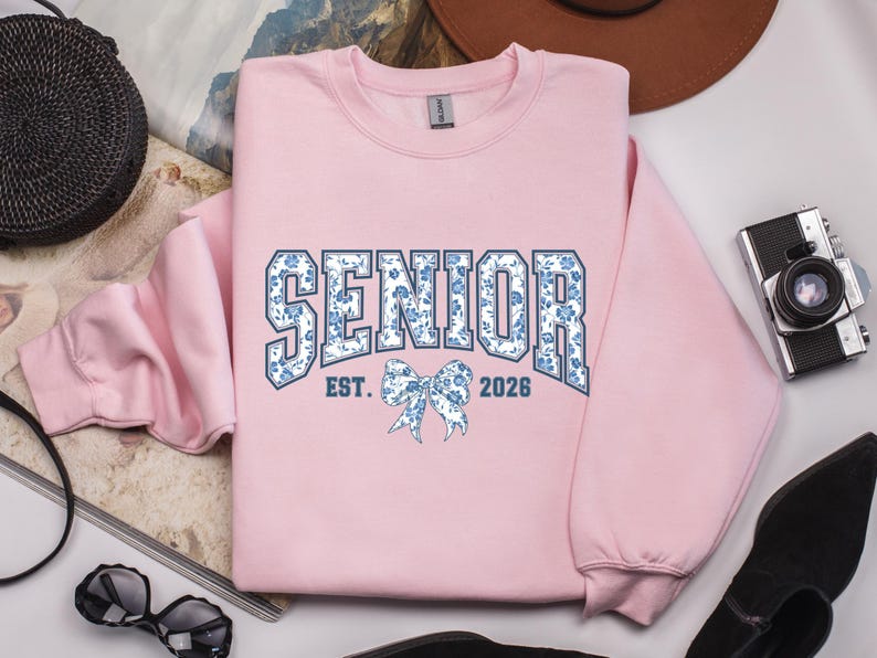 May include: A light pink sweatshirt with the word "SENIOR" in a blue floral design, with "EST. 2026" and a bow graphic. The sweatshirt is a crew neck style.