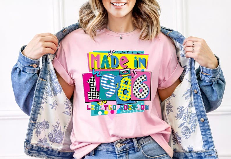 K&ouml;nnte beinhalten: Rosa T-Shirt mit einem farbenfrohen Grafikdesign mit der Aufschrift "MADE IN 1986 LIMITED EDITION". Das Design zeigt eine Kassette, Schachbrettmuster und Punkte. Das Shirt wird mit einer Jeansjacke getragen.