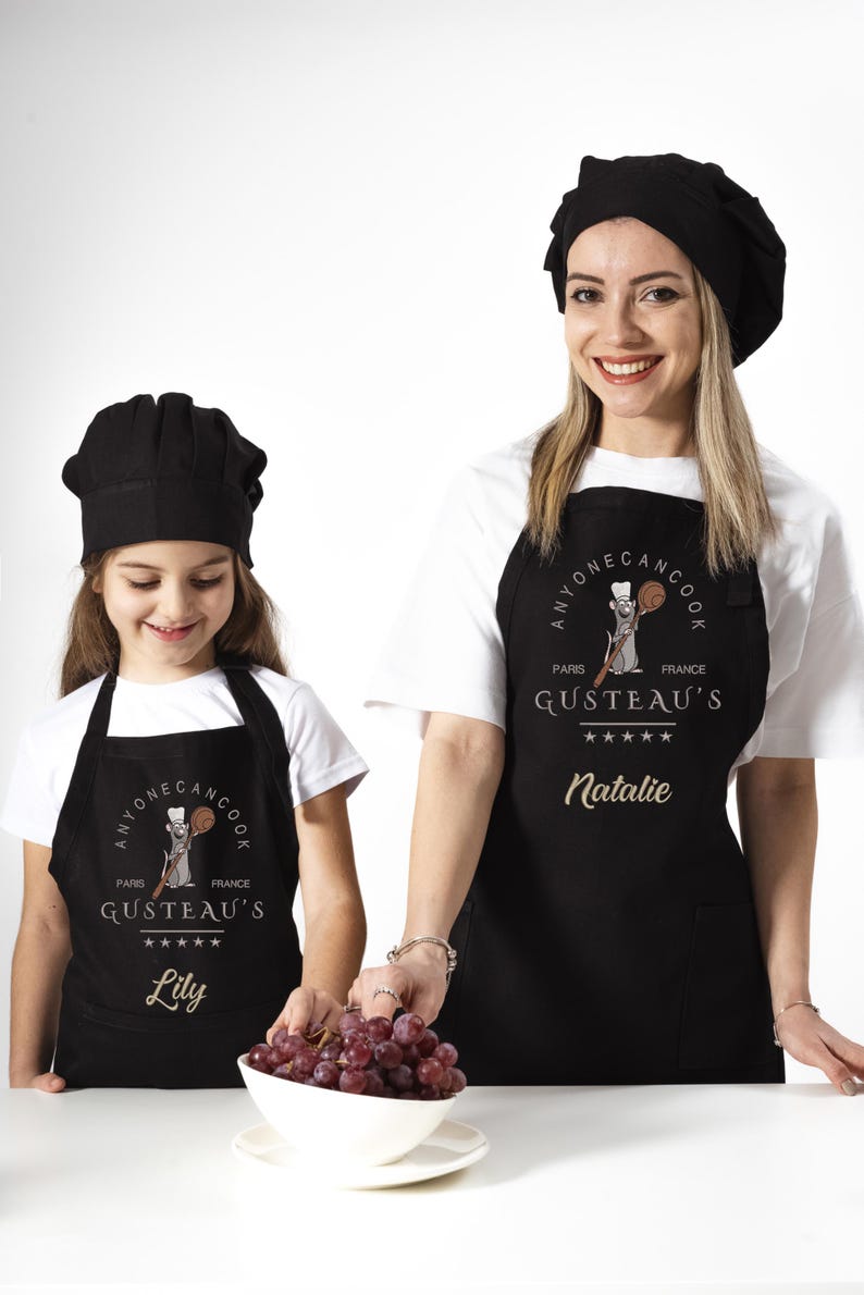 May include: Two people wearing black aprons with a cartoon rat chef design. The aprons say "Anyone Can Cook" and "Gusteau's" with the names "Natalie" and "Lily" printed below.