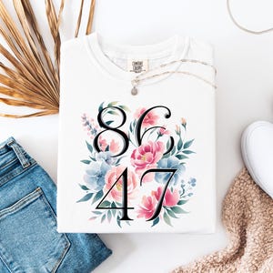 May include: White Comfort Colors sweatshirt with a floral design and the numbers 86 and 47. The floral arrangement includes pink and blue flowers with green leaves. A silver necklace and white sneakers are also visible.