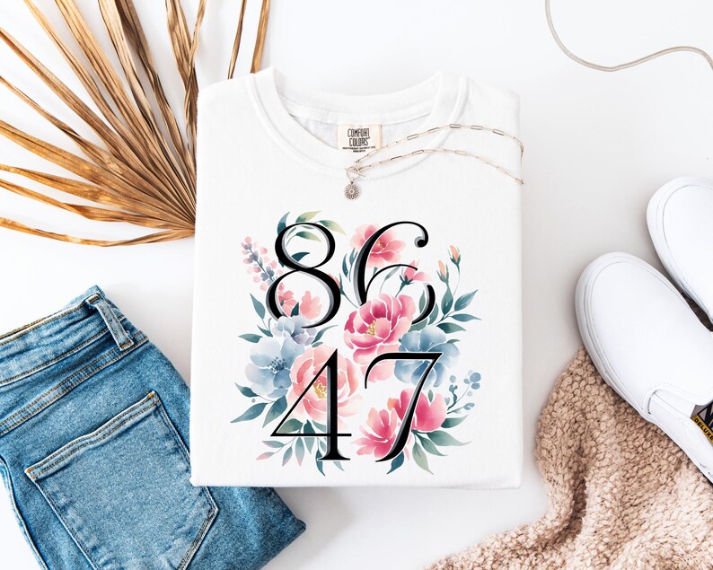 May include: White Comfort Colors sweatshirt with a floral design and the numbers 86 and 47. The floral arrangement includes pink and blue flowers with green leaves. A silver necklace and white sneakers are also visible.