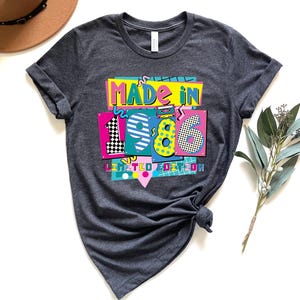 K&ouml;nnte beinhalten: Dunkelgraues T-Shirt mit einem farbenfrohen Aufdruck "MADE IN 1986 LIMITED EDITION". Das Design hat einen Retro-Look mit den Zahlen 1, 9, 8 und 6 in separaten Feldern. Das T-Shirt ist am unteren Ende geknotet.