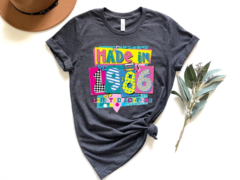 K&ouml;nnte beinhalten: Dunkelgraues T-Shirt mit einem farbenfrohen Aufdruck "MADE IN 1986 LIMITED EDITION". Das Design hat einen Retro-Look mit den Zahlen 1, 9, 8 und 6 in separaten Feldern. Das T-Shirt ist am unteren Ende geknotet.