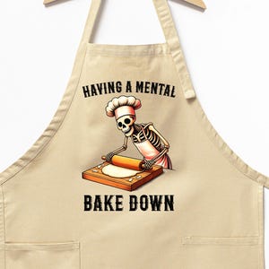 May include: A beige kitchen apron with a skeleton chef rolling out dough. The text on the apron reads "Having a mental bake down."