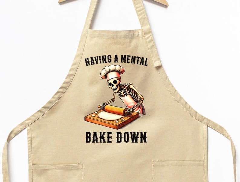 May include: A beige kitchen apron with a skeleton chef rolling out dough. The text on the apron reads "Having a mental bake down."
