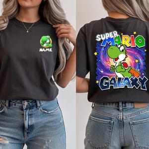 May include: Charcoal gray t-shirt featuring a green Yoshi character from Super Mario. The front has a small Yoshi graphic with the word "NAME" below. The back displays "SUPER MARIO GALAXY" with Yoshi in a galaxy-themed background.