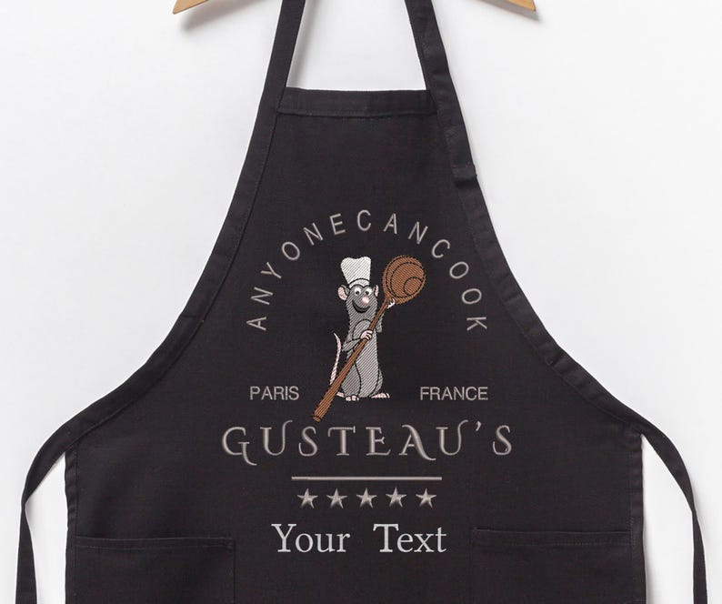 May include: Black kitchen apron with a white graphic of a rat chef holding a wooden spoon. The graphic includes the text "Anyone Can Cook", "Paris", "France", "Gusteau's", and five stars. The apron has a pocket and the text "Your Text" is printed below the graphic.