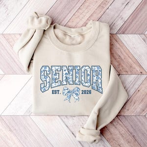 May include: A beige sweatshirt with the word "SENIOR" in blue floral print lettering. Below the word "SENIOR" is a blue floral bow and the text "EST. 2026". The sweatshirt has long sleeves and a crew neck.