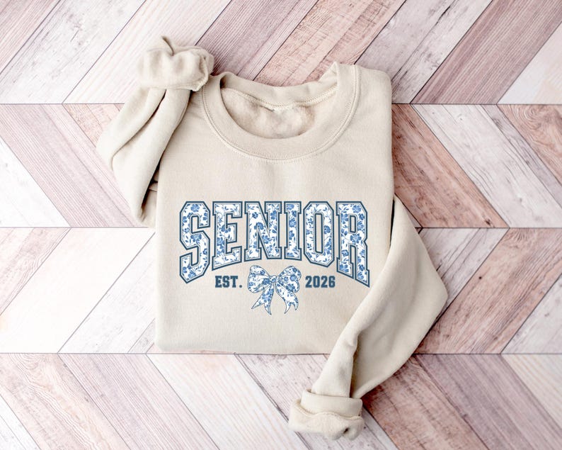 May include: A beige sweatshirt with the word "SENIOR" in blue floral print lettering. Below the word "SENIOR" is a blue floral bow and the text "EST. 2026". The sweatshirt has long sleeves and a crew neck.