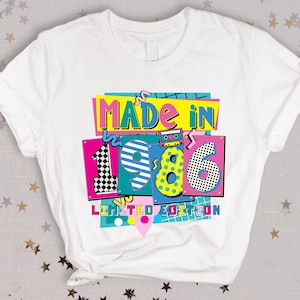 K&ouml;nnte beinhalten: Wei&szlig;es T-Shirt mit einem farbenfrohen Aufdruck mit der Aufschrift "MADE IN 1986 LIMITED EDITION". Das Design zeigt eine Retro-Kassette, Schachbrettmuster und leuchtende Farben wie Pink, Blau und Gelb. Ein lustiges T-Shirt im Vintage-Stil.
