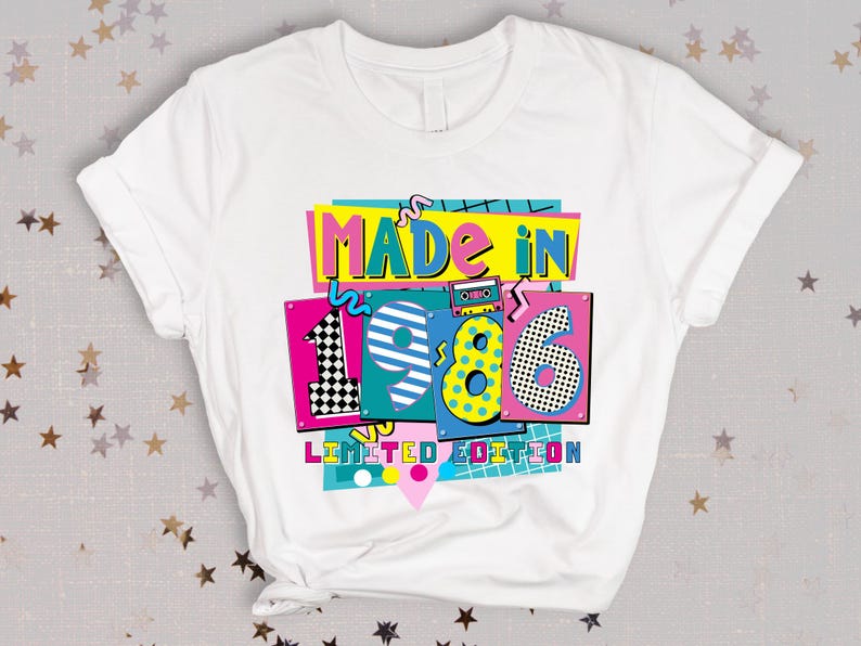 K&ouml;nnte beinhalten: Wei&szlig;es T-Shirt mit einem farbenfrohen Aufdruck mit der Aufschrift "MADE IN 1986 LIMITED EDITION". Das Design zeigt eine Retro-Kassette, Schachbrettmuster und leuchtende Farben wie Pink, Blau und Gelb. Ein lustiges T-Shirt im Vintage-Stil.
