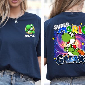 May include: Navy blue t-shirt featuring a green Yoshi character from Super Mario. The front displays Yoshi with the word "NAME" below. The back showcases Yoshi in a galaxy design with the words "SUPER MARIO GALAXY".