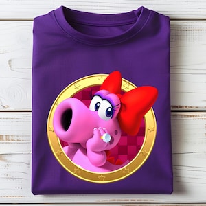 Peut inclure: Un t-shirt violet avec un personnage de dessin anim&eacute; rose, un n&oelig;ud rouge et une bague, dans un cercle dor&eacute;. Le t-shirt est pli&eacute; sur une surface en bois blanc. Le t-shirt est pli&eacute; sur une surface en bois blanc.