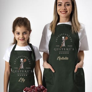 May include: Two green aprons with a cartoon rat chef design. The aprons have the text "Anyone Can Cook" and "Gusteau's" with a five-star rating. The aprons are personalized with the names "Lily" and "Natalie".