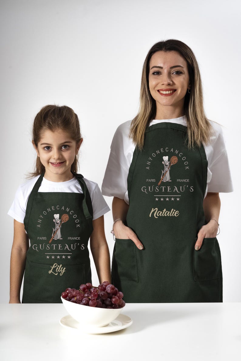May include: Two green aprons with a cartoon rat chef design. The aprons have the text "Anyone Can Cook" and "Gusteau's" with a five-star rating. The aprons are personalized with the names "Lily" and "Natalie".