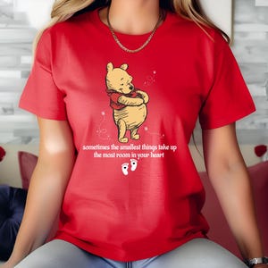 May include: Red t-shirt with a cartoon image of Winnie the Pooh hugging himself. The shirt has the text "sometimes the smallest things take up the most room in your heart" and two footprints below the image.