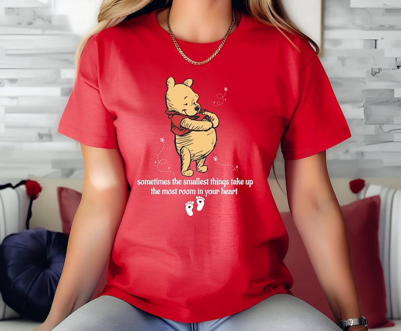 May include: Red t-shirt with a cartoon image of Winnie the Pooh hugging himself. The shirt has the text "sometimes the smallest things take up the most room in your heart" and two footprints below the image.