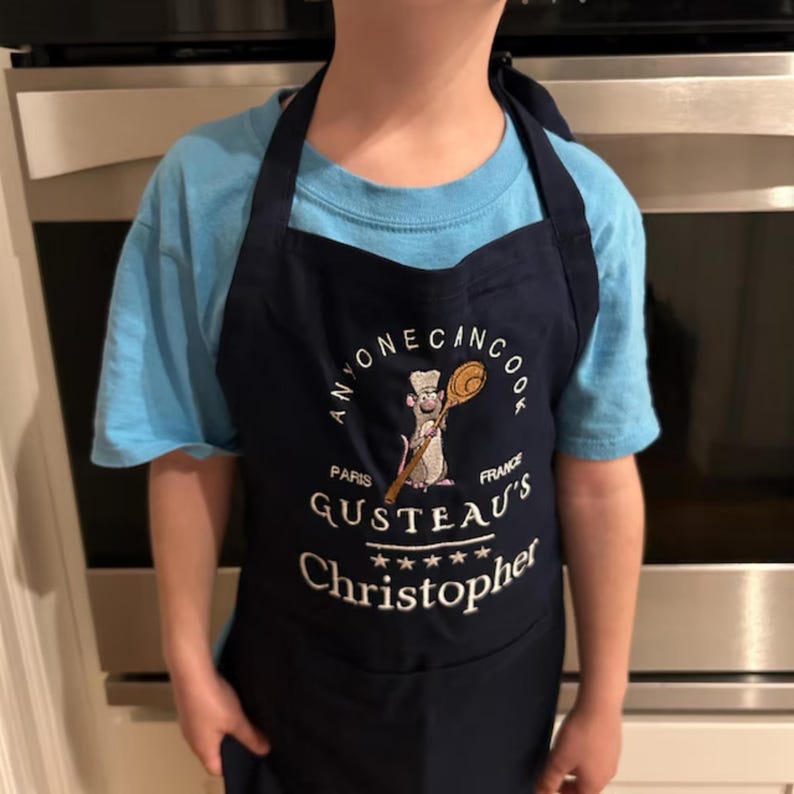 May include: A navy blue child's apron with a white embroidered design. The design features a cartoon rat chef holding a wooden spoon, the words "Anyone Can Cook", "Paris", "France", "Gusteau's", and five stars. The apron also has the name "Christopher" embroidered on it.