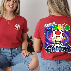 May include: Red t-shirt featuring a Super Mario Galaxy design. The front of the shirt has a small Toad graphic. The back of the shirt displays a larger graphic of Toad with the words "Super Mario Galaxy".