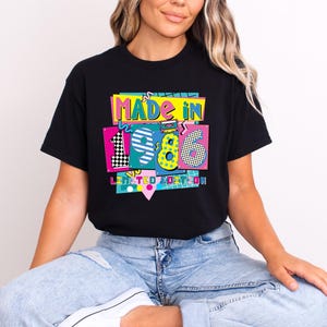 K&ouml;nnte beinhalten: Schwarzes T-Shirt mit einem farbenfrohen Grafikdesign mit der Aufschrift "MADE IN 1986 LIMITED EDITION". Das Design zeigt die Zahlen 1, 9 und 6 in verschiedenen Mustern und Farben im Retro-Stil der 80er Jahre. Das T-Shirt wird von einer Person getragen.