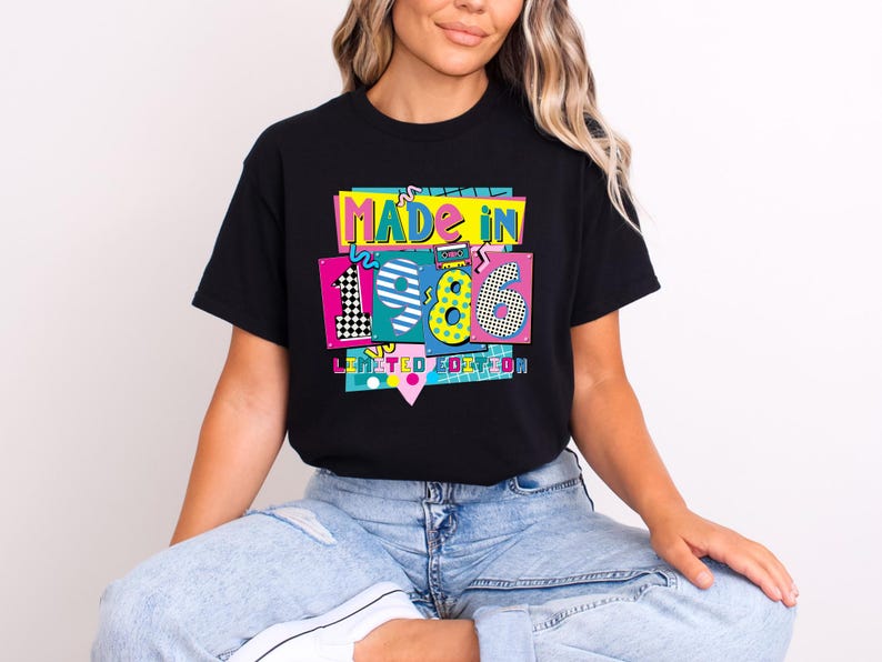 K&ouml;nnte beinhalten: Schwarzes T-Shirt mit einem farbenfrohen Grafikdesign mit der Aufschrift "MADE IN 1986 LIMITED EDITION". Das Design zeigt die Zahlen 1, 9 und 6 in verschiedenen Mustern und Farben im Retro-Stil der 80er Jahre. Das T-Shirt wird von einer Person getragen.