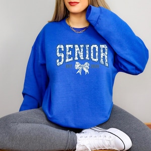 May include: A royal blue sweatshirt with the word "SENIOR" in a white floral design. The sweatshirt also features "EST 2026" and a bow graphic. The sweatshirt is worn with gray leggings and white sneakers.