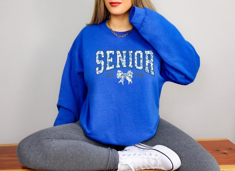 May include: A royal blue sweatshirt with the word "SENIOR" in a white floral design. The sweatshirt also features "EST 2026" and a bow graphic. The sweatshirt is worn with gray leggings and white sneakers.