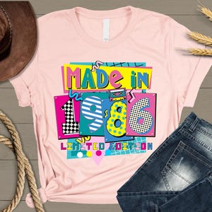 K&ouml;nnte beinhalten: Ein hellrosa T-Shirt mit dem Text "MADE IN 1986 LIMITED EDITION" in einem farbenfrohen Retro-Design. Die Jahreszahl "1986" ist mit verschiedenen Mustern und Farben hervorgehoben. Das Shirt wird mit einer blauen Jeans pr&auml;sentiert.